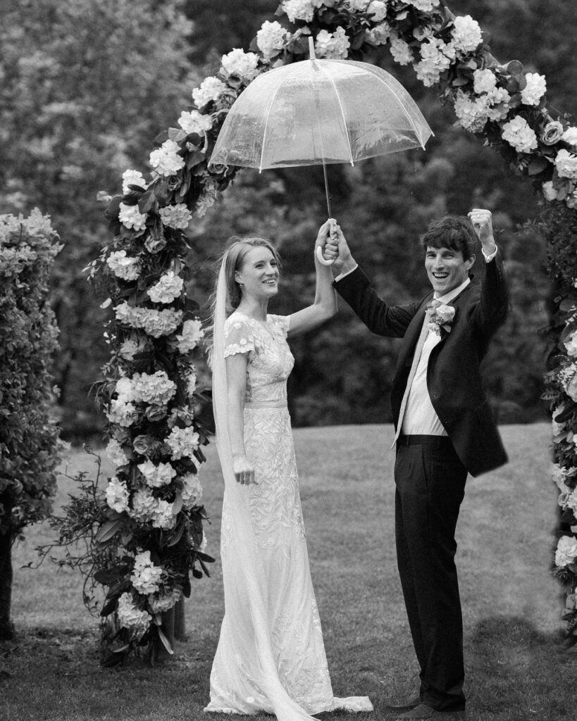 A delighted newly wed couple cheering, under an umbrella, during their humanist wedding ceremony