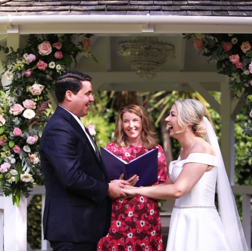 A wedding couple exchanging personalised wedding vows. They are laughing as their wedding celebrant looks on, smiling, as they make their promises. 