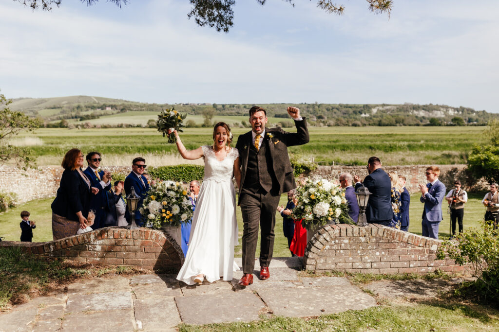 Newly weds cheering and punching the air with excitement as they reach the top of their wedding aisle. Their guests are cheering and clapping behind them.